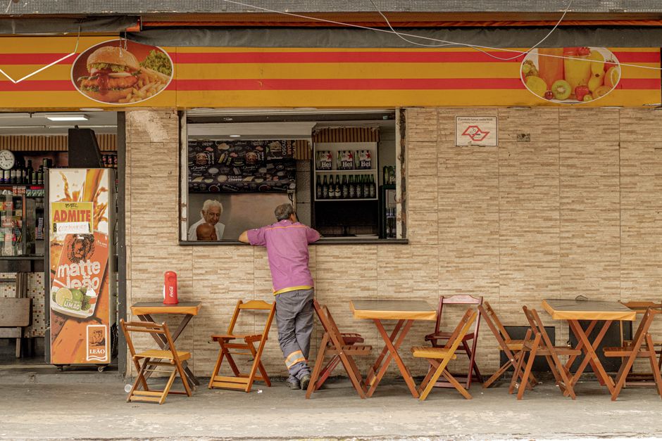 Uma pessoa vestindo uma camisa rosa está parada em frente a um café de rua com mesas e cadeiras de madeira.