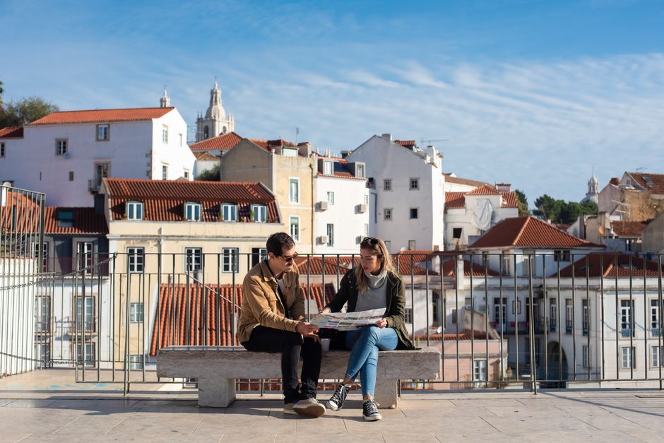 Um casal sentado em um banco olhando para um mapa com os telhados de Lisboa ao fundo.